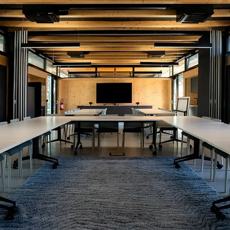 Sala riunioni con tavoli bianchi disposti a U, pareti e soffitto in legno e uno schermo sul fondo